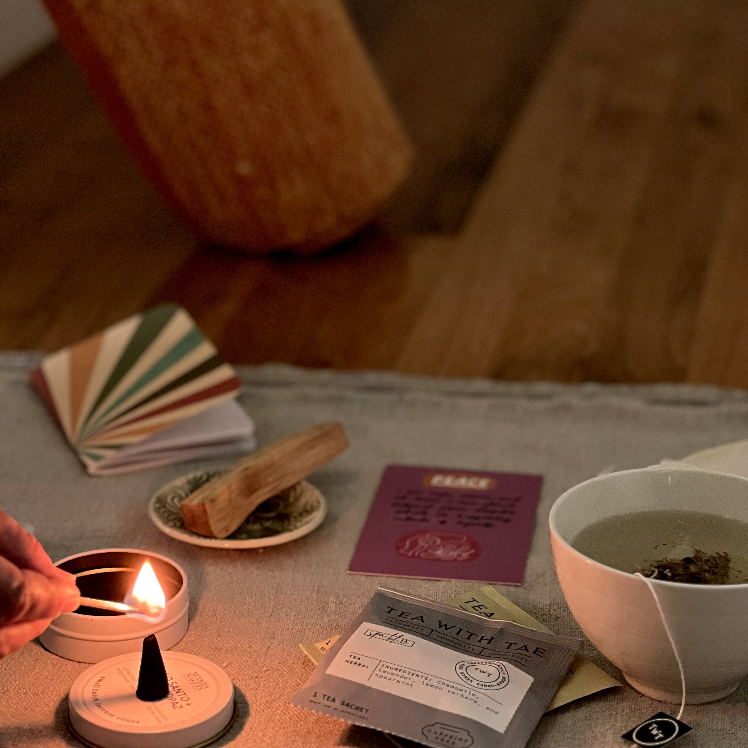 A cozy tabletop scene with a white teacup filled with tea and a tea bag, a lit candle, a small incense cone, a folded fan of colorful cards, a wooden piece, and a tea packet on a cloth-covered surface in a warm indoor setting.